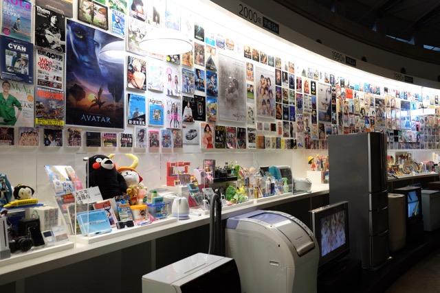 Long wall display of appliances, magazines, toys and posters from the 1960s through to the 2000s.